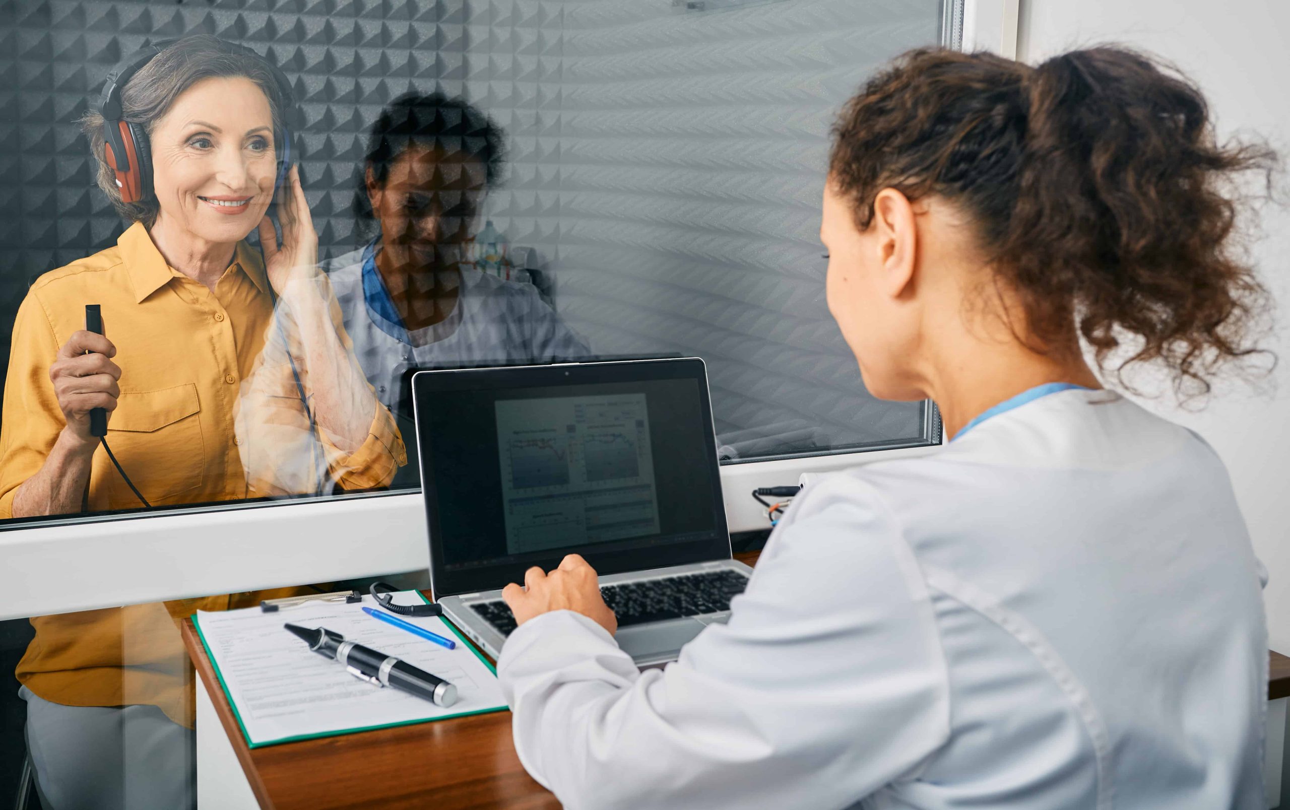 Women performing a hearing test inside a sound proof room. Another women operating hearing test machine. Close up.