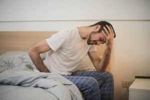 Man sitting on bed with hand placed on forehead.