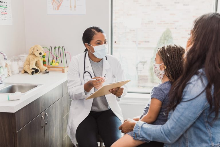 With a protective mask on, a female pediatrician talks to a young patient's mother about the woman's daughter's medical conditions. They are wearing protective masks during the COVID-19 pandemic.